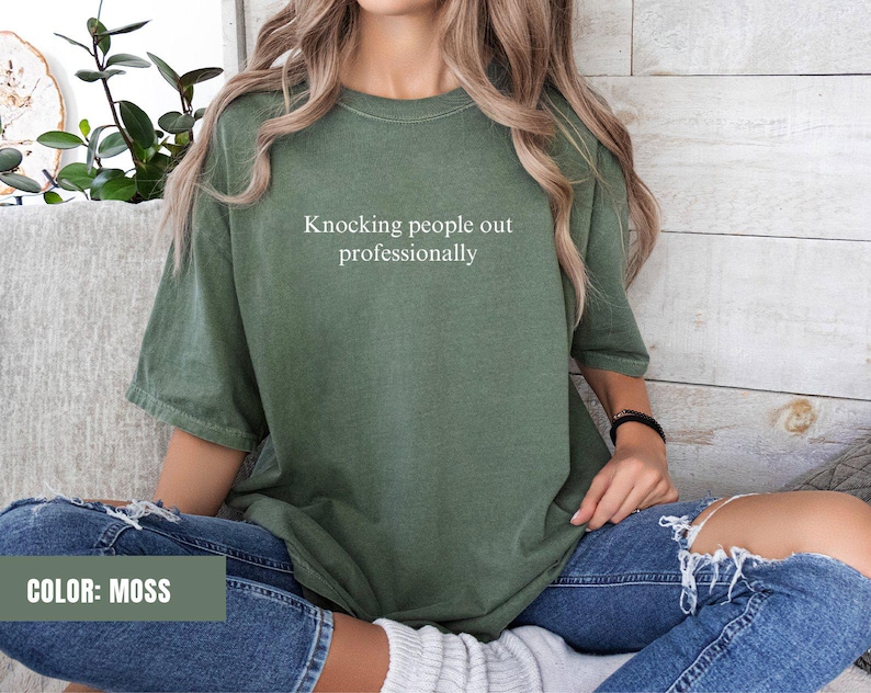 May include: A moss green t-shirt with the text "Knocking people out professionally" printed in white on the front. The shirt is being worn by a person with long brown hair and blue jeans.