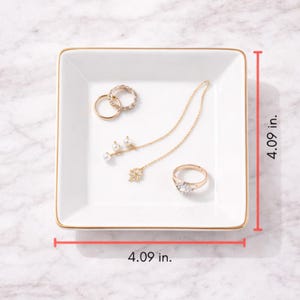 May include: A white square jewelry tray with gold trim, measuring 4.09 inches on each side. The tray holds several pieces of jewelry, including rings and a necklace with pearl accents. The background is a marble surface.