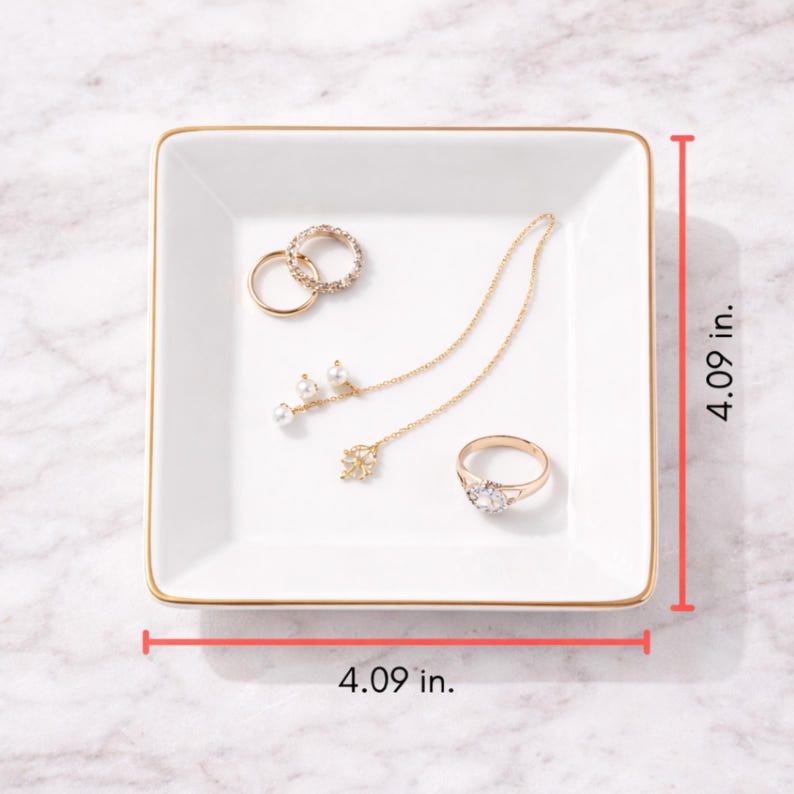 May include: A white square jewelry tray with gold trim, measuring 4.09 inches on each side. The tray holds several pieces of jewelry, including rings and a necklace with pearl accents. The background is a marble surface.