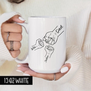 May include: White ceramic mug with a black design of three fists bumping each other. The text "Dad" is above the top fist, "Isla" is below the left fist, and "Jaden" is below the right fist. The mug is 15 ounces.