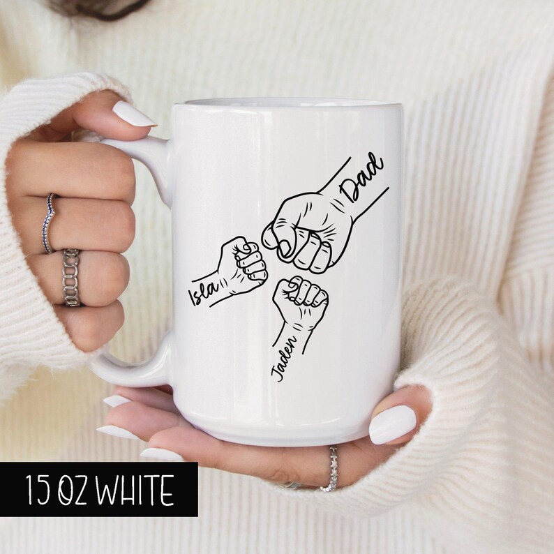 May include: White ceramic mug with a black design of three fists bumping each other. The text "Dad" is above the top fist, "Isla" is below the left fist, and "Jaden" is below the right fist. The mug is 15 ounces.