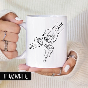 May include: White ceramic mug with a black outline of three fists bumping together. The text "Dad", "Isla", and "Jaden" are written on the fists. The mug is 11 ounces.