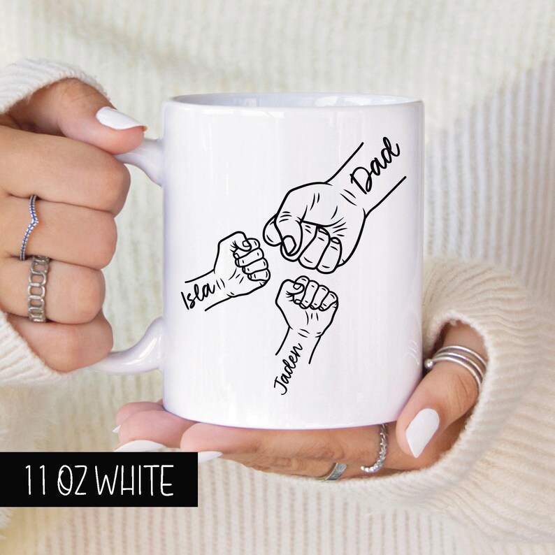 May include: White ceramic mug with a black outline of three fists bumping together. The text "Dad", "Isla", and "Jaden" are written on the fists. The mug is 11 ounces.