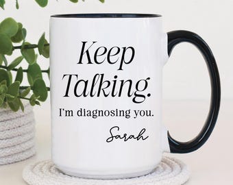 May include: White ceramic coffee mug with a black rim and handle. The mug has black text that reads "Keep Talking. I'm diagnosing you. Sarah".