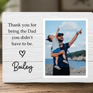 May include: White wooden picture frame with a photograph of a man and child pointing upwards. The frame has the text "Thank you for being the Dad you didn't have to be." and the name "Bailey".
