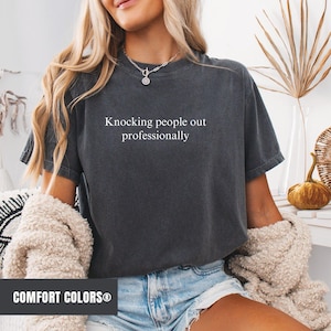 May include: A dark grey t-shirt with the text "Knocking people out professionally" printed in white. The shirt is made of a soft, comfortable material and has a relaxed fit.