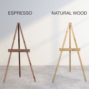 K&ouml;nnte beinhalten: Zwei Holzstaffeleien, eine in Espresso-Braun und eine in Naturholz. Beide Staffeleien haben ein schlichtes Design mit drei Beinen und einer horizontalen Stange zum Halten einer Leinwand oder eines Schilds.