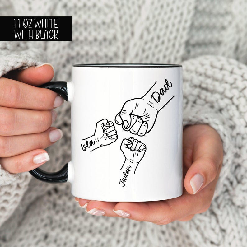 May include: White ceramic mug with a black rim and handle. The mug features a black and white illustration of three fists bumping together. The fists are labeled "Dad", "Isla", and "Jaden".