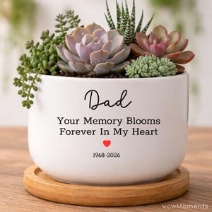 May include: A white ceramic planter with a variety of succulents, including green, pink, and purple hues. The planter has the word "Dad" in script, along with the message "Your Memory Blooms Forever In My Heart" and a red heart symbol. The years 1968-2026 are also included.
