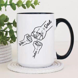 May include: White ceramic mug with a black rim and handle. The mug features a black and white illustration of three fists bumping together. The text "Dad" is written on the largest fist, and the text "Isla" and "Jaden" are written on the smaller fists.