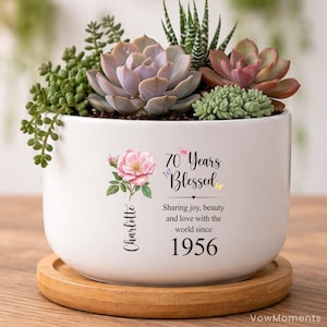 May include: A white ceramic planter on a round wooden base, filled with various green and pink succulents. The planter features a pink rose illustration, small butterflies, and the text: 70 Years Blessed, Charlotte, Sharing joy, beauty and love with the world since 1956.