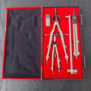 May include: A set of drawing instruments in a red case. The set includes a large compass, a small compass, and other tools. The case is open, showing the tools against a red background.