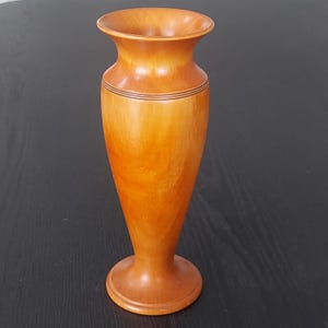 May include: A handcrafted wooden vase with a flared top and a rounded base. The vase is a warm, light brown colour with darker brown bands near the top. The vase is approximately 20 cm tall, perfect for displaying flowers or as a decorative piece.