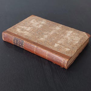 May include: An old book with a distressed, textured cover. The book features a brown leather spine and corners, with a lighter, speckled cover. The title is visible on the spine. The book is on a dark surface.