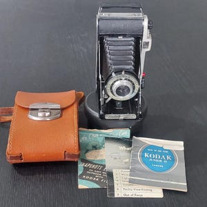 以下が含まれることがあります： 折りたたみ式のベローズデザインの、黒とシルバーのヴィンテージKodak Junior IIカメラ。茶色の革ケースといくつかの取扱説明書が含まれています。カメラは黒い台座の上にあります。
