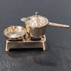 May include: A miniature gold-coloured toy kitchen set, including a pot with a lid, a frying pan, and a stove. The pot has a long handle, and the set is made of shiny metal. The stove has four legs.