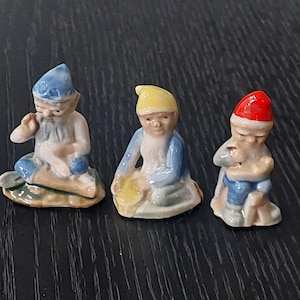 以下が含まれることがあります： 3つの小さなセラミック製のノームの置物。各ノームはひざまずいており、それぞれ異なる色の帽子をかぶっています。青、黄色、赤です。ノームは白いひげを生やし、青と白の服を着ています。緑色の台座の上にあります。