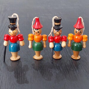 May include: Four wooden figurines of a soldier with painted details. Each figure has a red hat or top hat, red arms, blue or green skirt, and a walking stick. They are connected with a string for hanging.