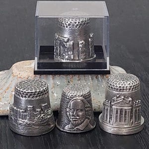 May include: A set of silver thimbles with intricate designs. One thimble is displayed in a clear case. The thimbles showcase detailed engravings of buildings and a portrait of William Shakespeare.