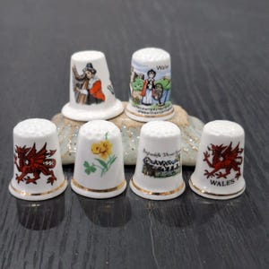 May include: A collection of six white ceramic thimbles featuring various colourful illustrations including a red dragon, a yellow daffodil, a person in traditional Welsh attire, and text reading Wales and Llanfairpwllgwyngyllgogerychwyrndrobwllllantysiliogogogoch.