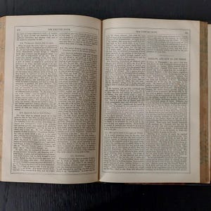 May include: An open book with aged, yellowed pages. The text is small and densely packed, with a title at the top of each page. The book is open on a dark surface, suggesting a vintage or antique item.
