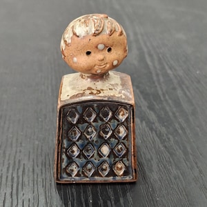 May include: A ceramic figurine featuring a stylised head with a tan complexion, white spots, and brown hair atop a rectangular base. The base displays a dark blue and brown diamond-patterned grid texture with a glossy finish and brown edges.