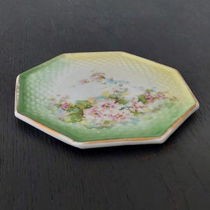May include: Octagonal porcelain plate with a green and yellow gradient background and a floral design featuring white and pink flowers. The plate has a gold rim.
