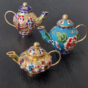 May include: Three miniature decorative teapots with floral designs. The teapots are gold, blue, and turquoise with colorful enamel flowers. Each teapot has a gold handle and spout. The teapots are displayed on a dark surface.
