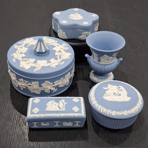 May include: A collection of five light blue jasperware pieces with white relief ornamentation. The set includes a round lidded box with grape motifs, a flower-shaped lidded box, a small rectangular lidded box, a round lidded box, and a pedestal vase.