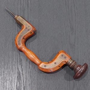 May include: A vintage hand drill with a wooden handle and a metal drill bit. The drill has brass and silver-coloured metal accents. The handle is curved and has a dark brown knob at the end. The tool is designed for woodworking.