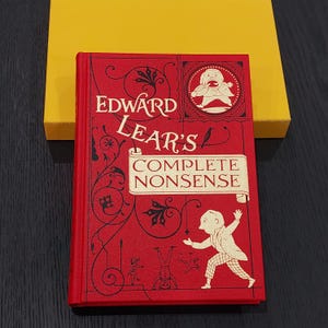 May include: A hardcover book titled "Edward Lear's Complete Nonsense" with a red cover and gold lettering. The book features whimsical illustrations and is set against a yellow box.