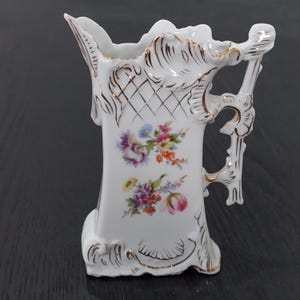 May include: White porcelain jug with a decorative handle and floral design. The jug features gold trim and a colourful bouquet of flowers. The handle has an ornate design. The jug is approximately 18 cm tall.