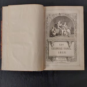 May include: Open antique book titled "The Leisure Hour" from 1855. The cover features an illustration of a family reading together. The book is open, revealing aged, cream-colored pages. The cover also includes the location "London" and the address "Paternoster Row, 164 Piccadilly."