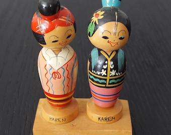 May include: Two vintage wooden dolls with painted details. One doll has a red and black hairstyle and a pink and white outfit. The other doll has a black hairstyle with a flower and a black and teal outfit. Both dolls are on a wooden base that says "KAREN".