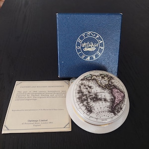 May include: A white porcelain globe with a detailed map of the Eastern and Western Hemispheres. The globe is in a blue box with the words "Optimago" and "World Paperweights" printed on it. The globe is a reproduction of a 19th century map.