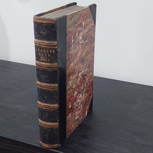 May include: An antique book titled "Leisure Hour 1855." The book has a dark brown leather spine with gold accents and text. The cover is a mix of red, brown, and cream colors. The book is standing upright on a dark surface.