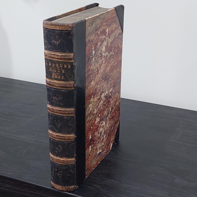 May include: An antique book titled "Leisure Hour 1855." The book has a dark brown leather spine with gold accents and text. The cover is a mix of red, brown, and cream colors. The book is standing upright on a dark surface.