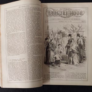 May include: An open book with a black and white illustration of a group of people in period clothing. The title "The Leisure Hour" is at the top, with text below. The page is from a historical publication.