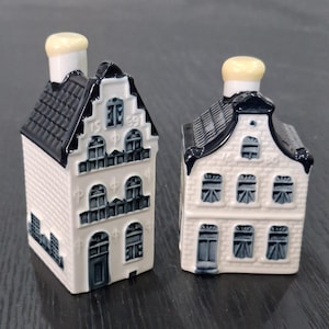 May include: Two ceramic miniature house figurines featuring a white facade with dark blue detailing. The taller house displays the year 1569 on its gable, while the shorter house shows 1680. Both structures have dark blue roofs and yellow chimneys.