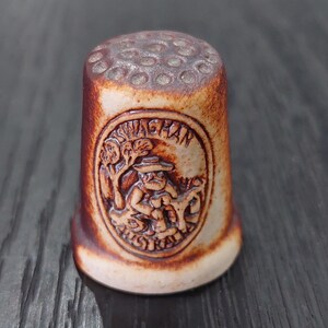 May include: A ceramic thimble with a brown and tan color scheme. The thimble features a detailed relief of a man, tree, and the word "Swagman Australia". The top of the thimble has small circular indentations.