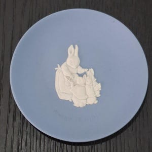 May include: A round, light blue ceramic plate featuring a raised white relief design of three rabbits. The central figure is a larger rabbit standing beside two smaller rabbits. The surface has a matte finish with a smooth, circular edge.