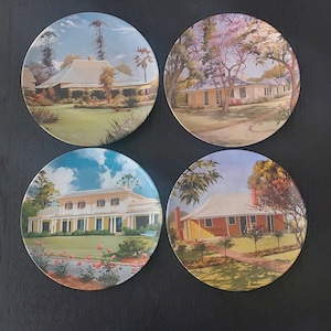 May include: Set of four ceramic plates featuring painted images of houses with green lawns and trees. Each plate has a different house design and colour scheme.