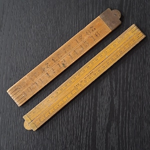 May include: Two vintage wooden folding rulers. One ruler is partially open, displaying markings in inches from 1 to 25. The other ruler is folded, showing markings in inches from 10 to 20. Both rulers have metal end pieces.