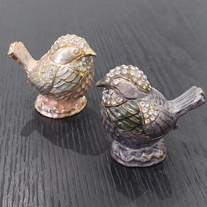May include: Two decorative bird figurines. The birds are silver and gold with crystal accents on their heads. The birds have detailed feather designs and are perched on small bases. The figurines are on a dark surface.
