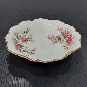 May include: A white, scalloped-edge porcelain dish with a floral rose pattern. The dish has a gold trim and is decorated with pink roses and green leaves. The dish is approximately 20 cm long and 13 cm wide.