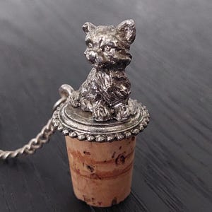 May include: A silver bottle stopper featuring a detailed cat figurine on top. The stopper is made of cork and has a silver chain attached.