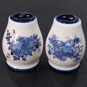 May include: Two white ceramic salt and pepper shakers with a blue floral pattern. Each shaker has a dark blue rim at the top and base. The shakers are pear-shaped and are a classic design.
