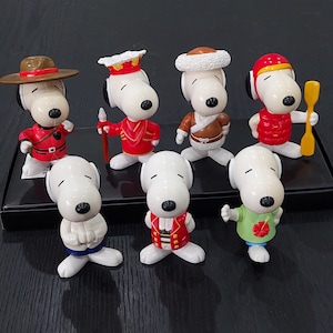 May include: A collection of Snoopy figurines in various outfits. The figures are white with black ears and noses. Outfits include a red uniform with a brown hat, a red and gold uniform, a brown fur-lined hat and coat, and a red outfit with a yellow paddle.