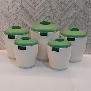 May include: A collection of five vintage kitchen canisters. The canisters are off-white with green lids and decorative green details. The canisters vary in size, creating a tiered arrangement.