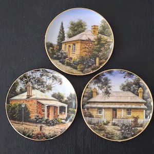 May include: Three round decorative plates with gold-toned rims, each featuring a detailed illustration of a stone cottage. The scenes depict various rural houses with chimneys, surrounded by trees, bushes, and garden landscapes in natural colours.